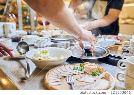 A hand reaches out to take a portion of a variety of dishes from the buffet, surrounded by many people enjoying a meal at a lively event. High quality photo A hand reaches out to take a portion of a variety of dishes from the buffet, surrounded by many people enjoying a meal at a lively event. High quality photo 121933946