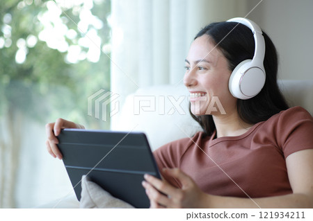 Happy asian woman listening audio holding tablet looking away 121934211
