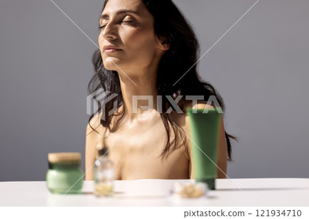 Close-up studio portrait of attractive young woman with her eyes closed in sunlight. Jars of cream and serum in foreground. Care about clean and soft face skin. 121934710