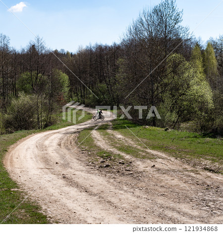 Cyclists on a winding dirt road through a forest. A sunny day with trees and greenery. Cyclists on a winding dirt road through a forest. A sunny day with trees and greenery. 121934868