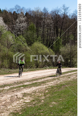 Two cyclists enjoy a sunny ride on a dirt road through a forest. Spring foliage provides a vibrant backdrop. Two cyclists enjoy a sunny ride on a dirt road through a forest. Spring foliage provides a vibrant backdrop. 121934869