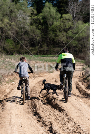Father and son cycling on a dirt road with their dog. 121934885