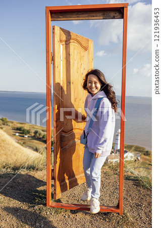 A young attractive Asian woman laughs and opens the door outdoors. A door on a hill and a beautiful seascape. 121936453