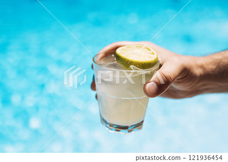 Young male hand holds a glass of a chilled drink with ice cubes. White cocktail with a lemon slice. Sunny day and reflection of rays in the blue pool water on background. 121936454