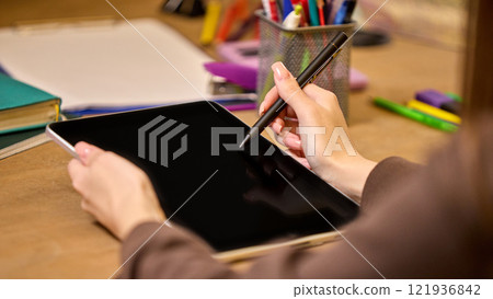 Back view of woman using tablet with stylus, surrounded by notebooks, colorful stationery, and tidy desk, symbolizing focus and productivity. Back view of woman using tablet with stylus, surrounded by notebooks, colorful stationery, and tidy desk, symbolizing focus and productivity. 121936842