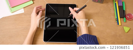Woman hands holding stylus while using tablet on wooden desk surrounded by colorful notebooks and office supplies, illustrating creativity and digital work. 121936851