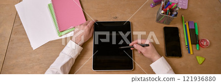 Person using stylus on digital tablet at desk surrounded by colorful stationery, notebooks, and smartphone. Top view 121936880