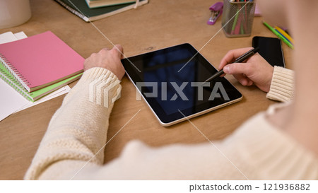 Close-up of person using stylus to write on digital tablet placed on a wooden desk, surrounded by notebooks and stationery. 121936882