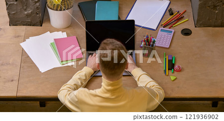 Overhead view of desk setup with person working on laptop, surrounded by colorful stationery and notebooks. 121936902