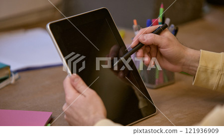 Close-up of hands holding tablet and stylus pointing at blank screen with organized desk setup, featuring colorful stationery, notebooks 121936909