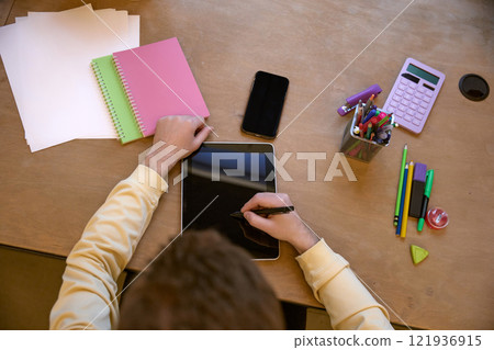 Close-up shot of hands using a stylus on a tablet in a workspace filled with colorful notebooks, pens, and a calculator. 121936915