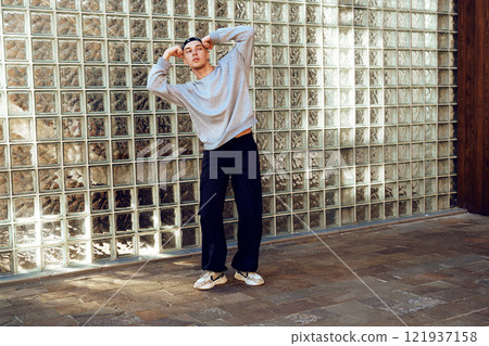 A young man wearing a gray sweater and black pants stands in front of a glass block wall, posing with one arm raised above his head A young man wearing a gray sweater and black pants stands in front of a glass block wall, posing with one arm raised above his head 121937158