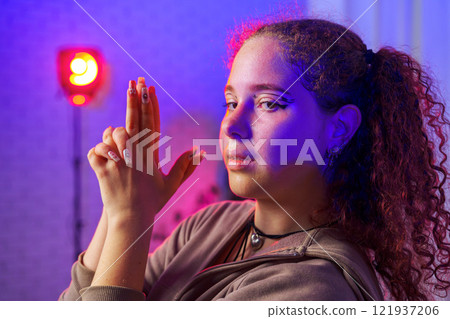 A young woman with curly hair makes a hand gesture, illuminated by a red light in a studio 121937206