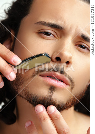 Man with curly hair grooming his beard carefully using a straight razor against a plain white background Man with curly hair grooming his beard carefully using a straight razor against a plain white background 121937313