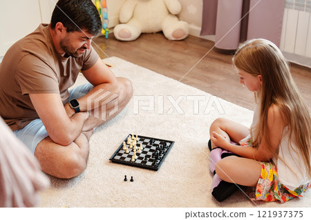 A father and daughter play chess on a white rug in a home 121937375
