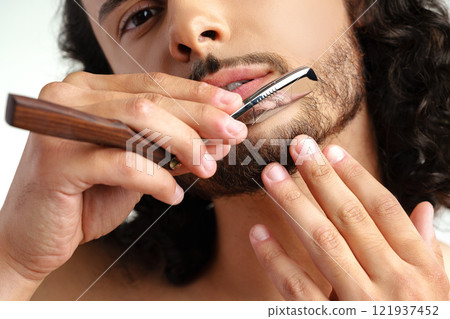 Man with curly hair grooming his beard carefully using a straight razor against a plain white background Man with curly hair grooming his beard carefully using a straight razor against a plain white background 121937452