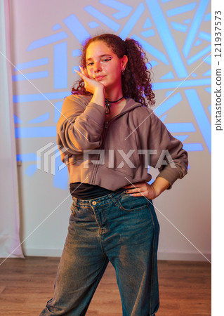 A young woman with curly hair poses in a dance studio, wearing a brown hoodie and blue jeans 121937573