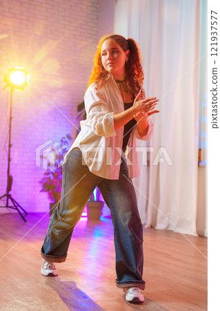 A young woman with curly red hair dances in a studio, wearing a white shirt and blue jeans A young woman with curly red hair dances in a studio, wearing a white shirt and blue jeans 121937577