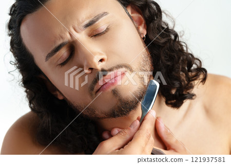 Man with curly hair grooming his beard carefully using a straight razor against a plain white background 121937581