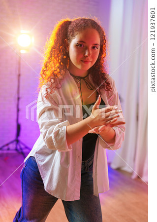 A young woman with curly red hair practices a dance move in a studio with purple lighting 121937601