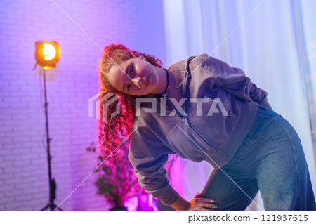 A young woman with pink hair poses in a studio, lit by a bright light 121937615