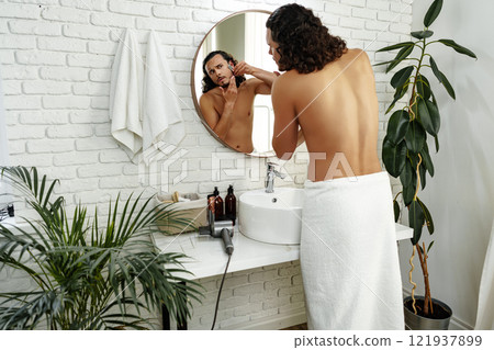 A man with curly hair grooming himself in the bathroom while looking into a round mirror A man with curly hair grooming himself in the bathroom while looking into a round mirror 121937899