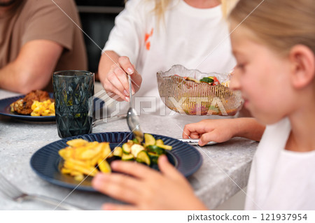 A young girl eats a meal of potatoes and vegetables while sitting at a table with family 121937954