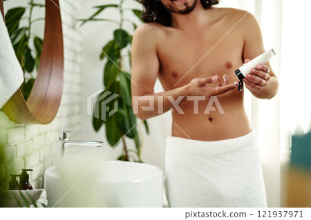 A young man applying skincare cream on his face with a focused expression, demonstrating personal grooming at home 121937971