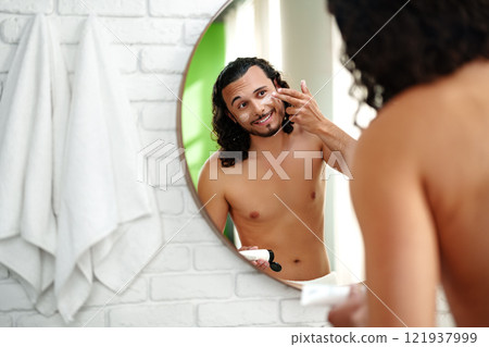 A young man applies skincare cream while looking in a round mirror in a bright bathroom A young man applies skincare cream while looking in a round mirror in a bright bathroom 121937999