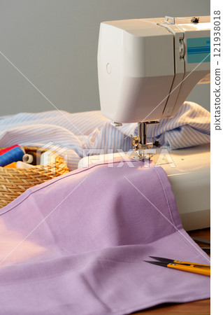 Sewing machine in use on a wooden table, stitching a light fabric in a well-lit indoor workspace Sewing machine in use on a wooden table, stitching a light fabric in a well-lit indoor workspace 121938018