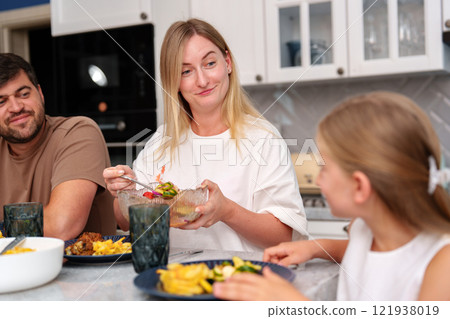 A woman sits at a kitchen table with her family, enjoying a meal together 121938019