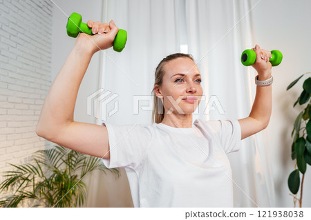 A woman exercises with green dumbbells at home in a bright room 121938038
