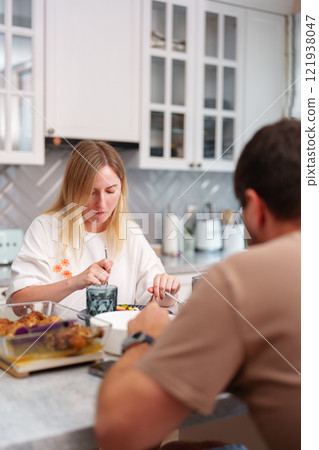 A woman eats a meal in her kitchen, while a man sits next to her A woman eats a meal in her kitchen, while a man sits next to her 121938047