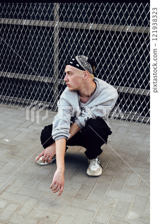 A young man wearing a bandana and casual clothes is squatting on the sidewalk in front of a chain-link fence A young man wearing a bandana and casual clothes is squatting on the sidewalk in front of a chain-link fence 121938323