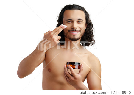 A young man applying skincare cream on his face with a focused expression, demonstrating personal grooming at home 121938960