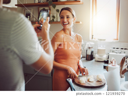 Woman cooking with husband taking a picture in kitchen on his mobile phone at home. Beautiful, happy and female chef cutting vegetables to cook healthy, delicious and vegetarian food at a table. Woman cooking with husband taking a picture in kitchen on his mobile phone at home. Beautiful, happy and female chef cutting vegetables to cook healthy, delicious and vegetarian food at a table. 121939270