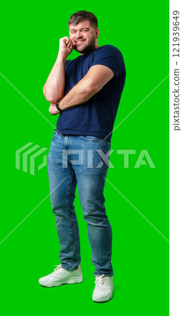 A man in a blue shirt and jeans poses confidently with a friendly smile against a bright green backdrop A man in a blue shirt and jeans poses confidently with a friendly smile against a bright green backdrop 121939649