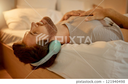 Happy man, streaming a podcast in bed smiling with headphones at home. Young guy listening to music from spotify on internet or radio in earphones, time to relax, enjoy peace in his apartment bedroom Happy man, streaming a podcast in bed smiling with headphones at home. Young guy listening to music from spotify on internet or radio in earphones, time to relax, enjoy peace in his apartment bedroom 121940498