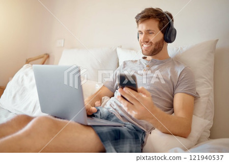 5g internet, video and online streaming of a man on a computer and phone in bed. Happy guy with a smile relax and multitask listening to a digital radio podcast or web music with modern technology 5g internet, video and online streaming of a man on a computer and phone in bed. Happy guy with a smile relax and multitask listening to a digital radio podcast or web music with modern technology 121940537