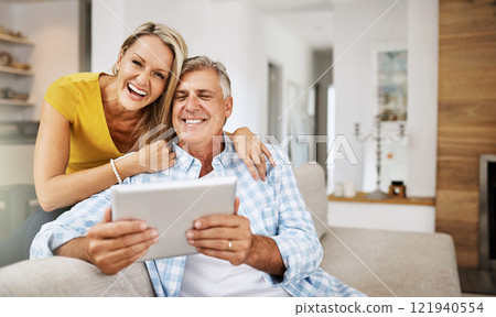 Mature couple laughing and smiling at funny app on digital tablet together, on their modern living room couch. Husband and wife sitting at home and looking at their touchscreen display device 121940554