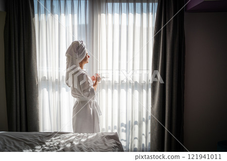 Beautiful young woman in a white bathrobe with a towel on her head stands near the window. 121941011