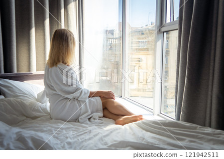 young woman in bathrobe sitting on bed and looking out the window 121942511