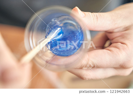 Close up of hands mixing blue epoxy resin in clear plastic cup with wooden Close up of hands mixing blue epoxy resin in clear plastic cup with wooden 121942663