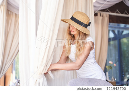 Caucasian female in sun hat adjusts curtains in bright room setting 121942700