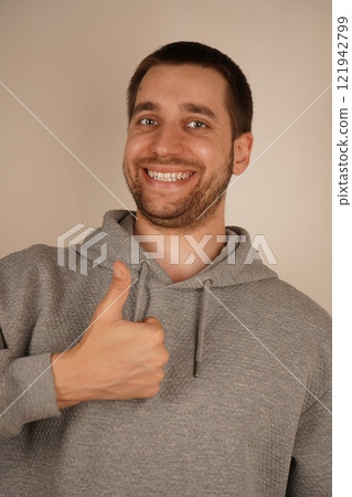 Young handsome man with beard over isolated gray background giving a thumbs up gesture. Approving expression looking at the camera. Charming confident guy dressed gray hoodie showing thumbs up alone. 121942799