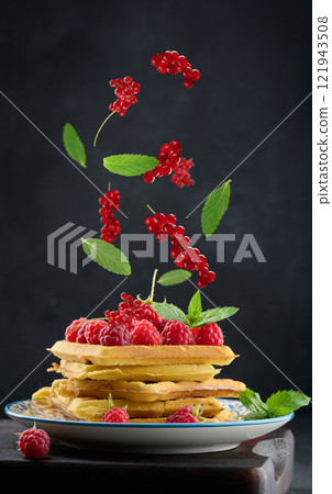 Stack of belgian waffles in a plate with raspberries and red currants on a black background, green mint leaves levitate Stack of belgian waffles in a plate with raspberries and red currants on a black background, green mint leaves levitate 121943508