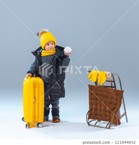Child in warm outfit holding snowball, standing near yellow suitcase and sled with gloves against grey studio background. 121944652