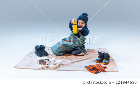 Curious little boy, sitting on floor and put vacuum flask into traveler backpack against gray studio background, after rest. 121944656