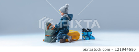 Banner. Curious little boy, dressed warm clothes, sitting on luggage ready to rest against grey studio background with negative space to insert text. 121944680