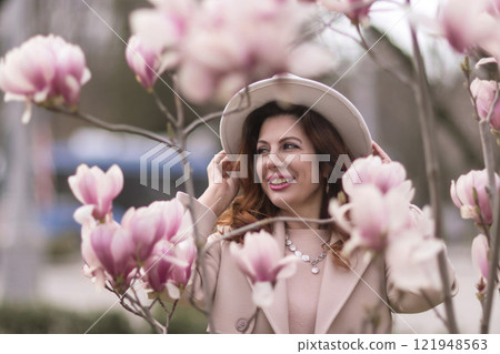 Woman magnolia flowers, surrounded by blossoming trees., hair down, white hat, wearing a light coat. Captured during spring, showcasing natural beauty and seasonal change. Woman magnolia flowers, surrounded by blossoming trees., hair down, white hat, wearing a light coat. Captured during spring, showcasing natural beauty and seasonal change. 121948563